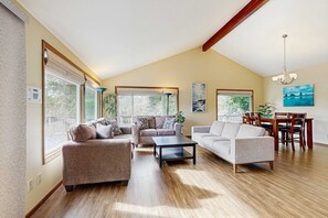 A spacious living room with a high ceiling, large windows, and a dining area with wooden furniture. - Charming riverfront home with deck, WiFi, streaming, W&D - 7 blocks to the beach (Seaside)