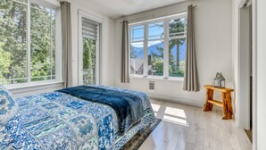 Quarto com cama grande, mesa de cabeceira de madeira e janela com vista para árvores e montanhas.