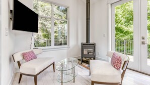 Uma sala de estar moderna com uma mesa de vidro, dois sofás brancos com almofadas estampadas, uma TV fixada na parede e uma lareira a lenha.