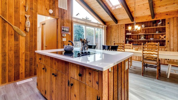 A wooden cabin interior with a kitchen island, dining area, and large windows.