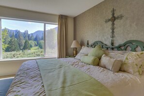 A bedroom with a bed, a cross on the wall, and a view of a forest through a window.