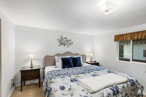 Une chambre à coucher avec un lit, deux tables de chevet avec des lampes, un élément décoratif mural et une fenêtre agrémentée d’une lambrequin.