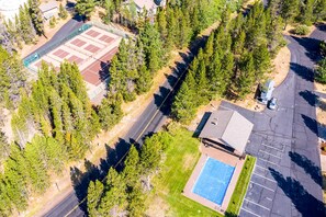 Vista aérea de una propiedad con cancha de tenis, piscina y zona de estacionamiento rodeada de árboles.