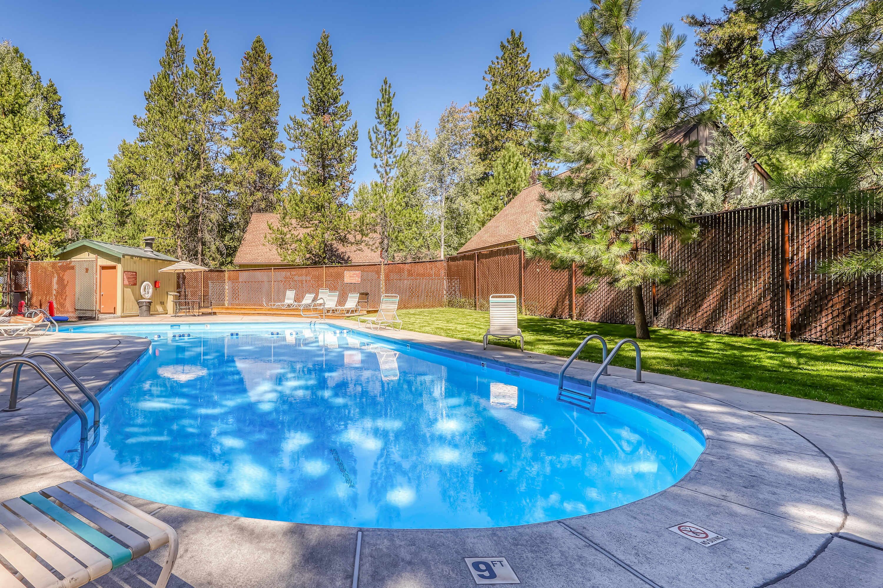 A swimming pool with a wooden deck, lounge chairs, and a fenced area with trees.