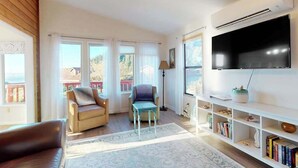 Sala de estar con sofá, sillón, televisor, estantería y vistas amplias por las ventanas.