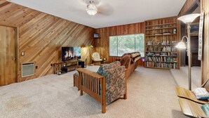 A cozy room with a wooden ceiling, a fireplace, a sofa, a bookshelf, and a window. - Spacious home with full kitchen, wifi, private backyard & grill (Welches)