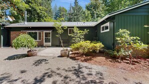 A house with a gravel driveway, a small front yard with potted plants, and a green building with a metal roof. - Spacious home with full kitchen, wifi, private backyard & grill (Welches)