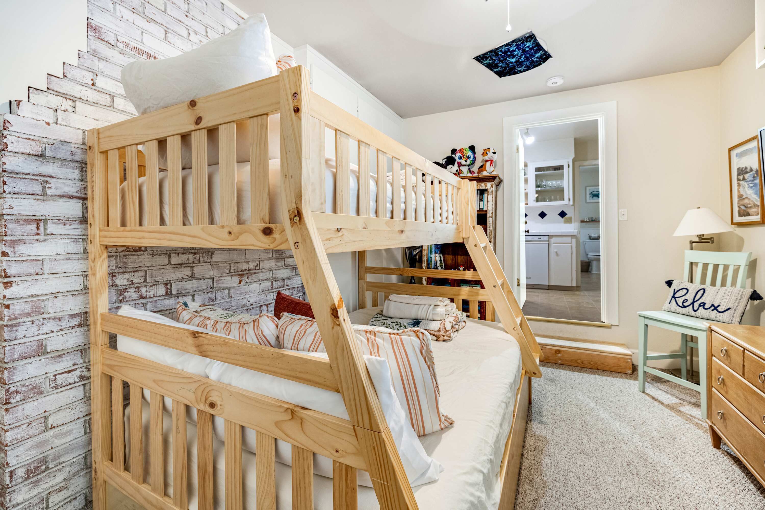 Un lit superposé avec une structure en bois, un matelas blanc et des oreillers à rayures. On aperçoit une petite table avec une lampe et une chaise avec un coussin portant l’inscription « Détendez-vous ». Un mur en briques et une porte donnant sur une salle de bain sont visibles.