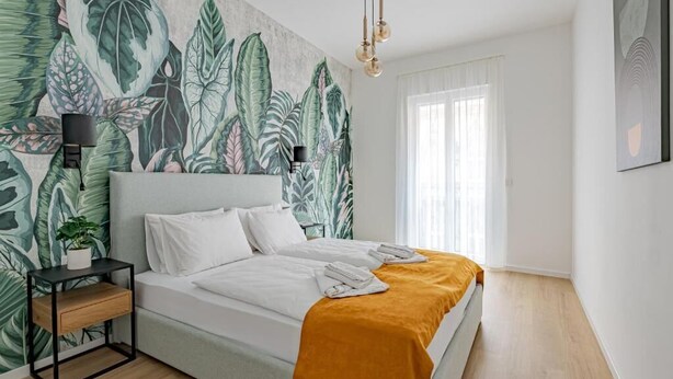 A bedroom with a bed, a nightstand, a lamp, a potted plant, and a large window. - D52 Apartment Hotel (Budapest)