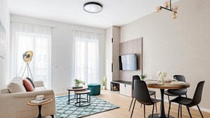 A modern living room with a sofa, coffee table, dining table with chairs, a TV mounted on the wall, and a floor lamp. - D52 Apartment Hotel (Budapest)
