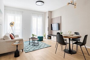 A modern living room with a sofa, coffee table, dining table with chairs, a TV mounted on the wall, and a floor lamp. - D52 Apartment Hotel (Budapest)