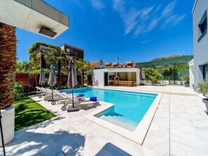 A modern outdoor pool area with lounge chairs, a barbecue, and a view of a mountain. - 5-bedroom Estate With Heated Pool (Kastela)