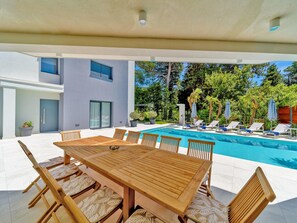A modern outdoor pool area with a wooden table and chairs, surrounded by a pool, potted plants, and a view of a building. - 5-bedroom Estate With Heated Pool (Kastela)