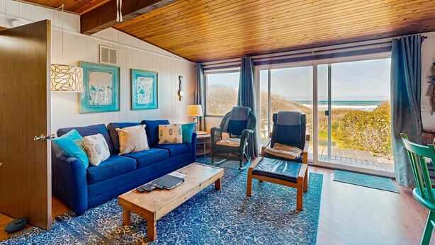 A room with a blue sofa, a wooden coffee table, a wicker chair, and a view of the ocean through large windows.