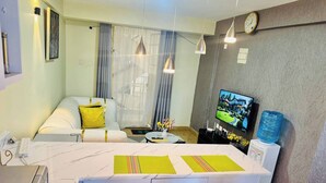 Quarto de hotel moderno com sofá, mesa de jantar com cadeiras, televisão e dispensador de água.