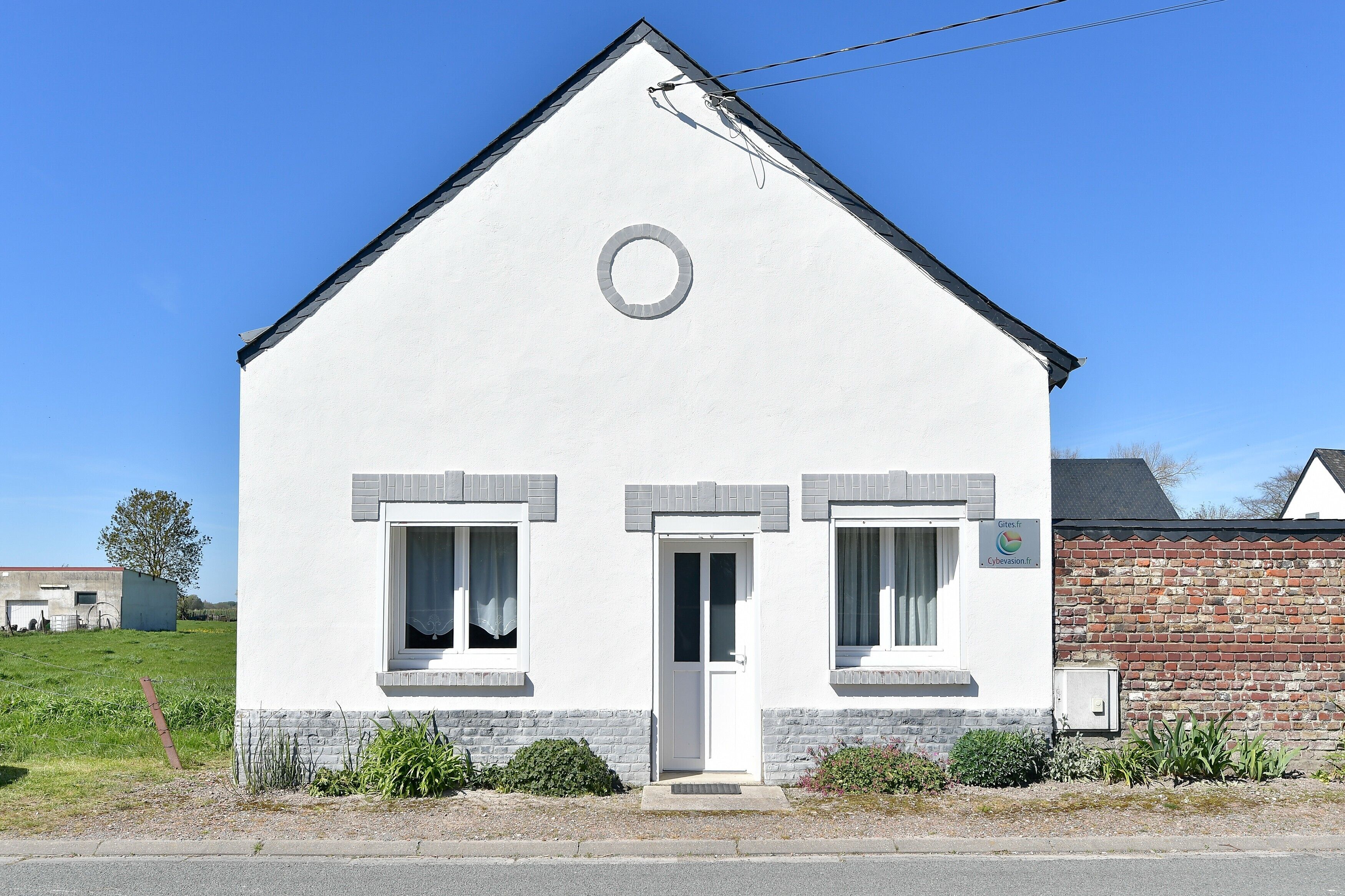 A white building with a gabled roof, two windows with curtains, a door, and a circular window above the door.