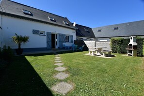 Ein weißes Gebäude mit einem geneigten Dach, einer gepflasterten Terrasse, Sitzgelegenheiten im Freien und einem Garten.