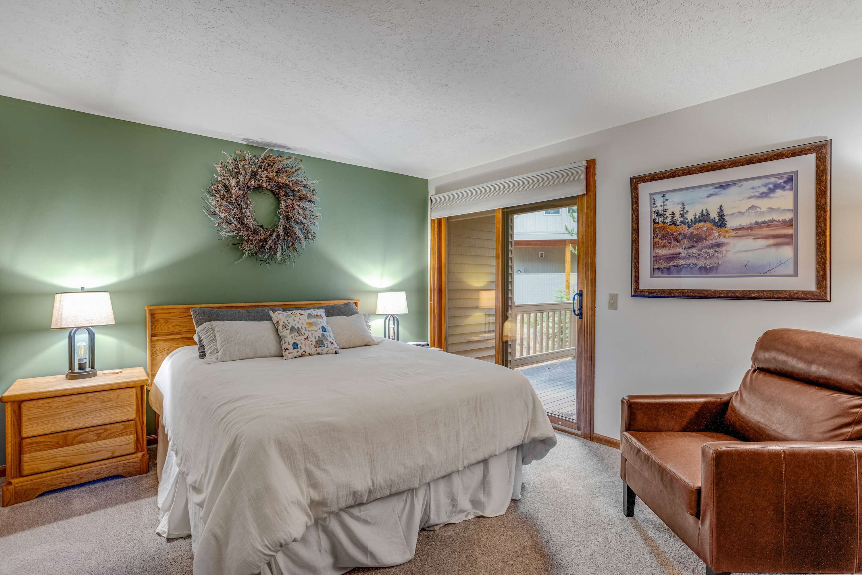A bedroom with a bed, nightstands, a brown leather chair, a wooden headboard, a green wall, a framed picture, and a view to the outside.