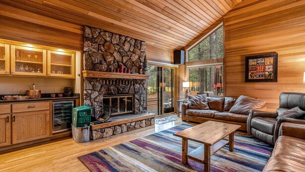 A cozy living room with a stone fireplace, leather sofas, and a large window offering a forest view. - Forested getaway with kitchen, stone fireplace, pool, & private hot tub (Black Butte Ranch)