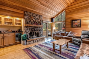A cozy living room with a stone fireplace, leather sofas, and a large window offering a forest view.