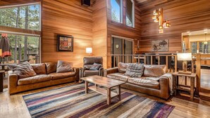 A spacious living room with leather sofas, a coffee table, and a large window. - Forested getaway with kitchen, stone fireplace, pool, & private hot tub (Black Butte Ranch)