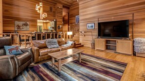 A spacious living room with leather sofas, a wooden coffee table, a flat-screen TV, and a staircase leading to an upper level. - Forested getaway with kitchen, stone fireplace, pool, & private hot tub (Black Butte Ranch)