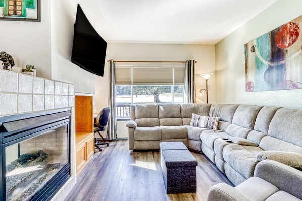 A living room with a sectional sofa, a fireplace, a TV mounted on the wall, and a window with blinds. - Two-story townhome on Ridge Golf Course with pools, hot tubs, gyms, tennis (Eagle Crest)