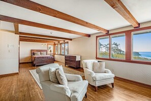 A spacious room with wooden beams, a large bed, two white armchairs, a dresser, and a view of the ocean.
