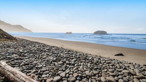 A pebbled beach with a log, ocean waves, and distant land. - Secluded beachfront home with Pacific Ocean views, fireplace, game room, grill (Arch Cape)