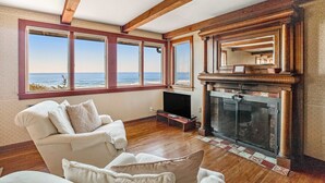 A living room with a fireplace, a large window with a view of the sea, and a wooden ceiling. - Secluded beachfront home with Pacific Ocean views, fireplace, game room, grill (Arch Cape)