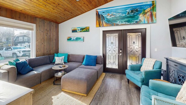 Ein modernes Wohnzimmer mit Holzboden, einem großen Fenster, einem Ecksofa, einem türkisfarbenen Sessel und einem Kamin.