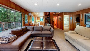 Ein geräumiges Wohnzimmer mit einer großen Eckcouch, einem Lederhocker und einer beigen Couch. Eine Holztreppe führt zu einem Essbereich mit Blick ins Freie.