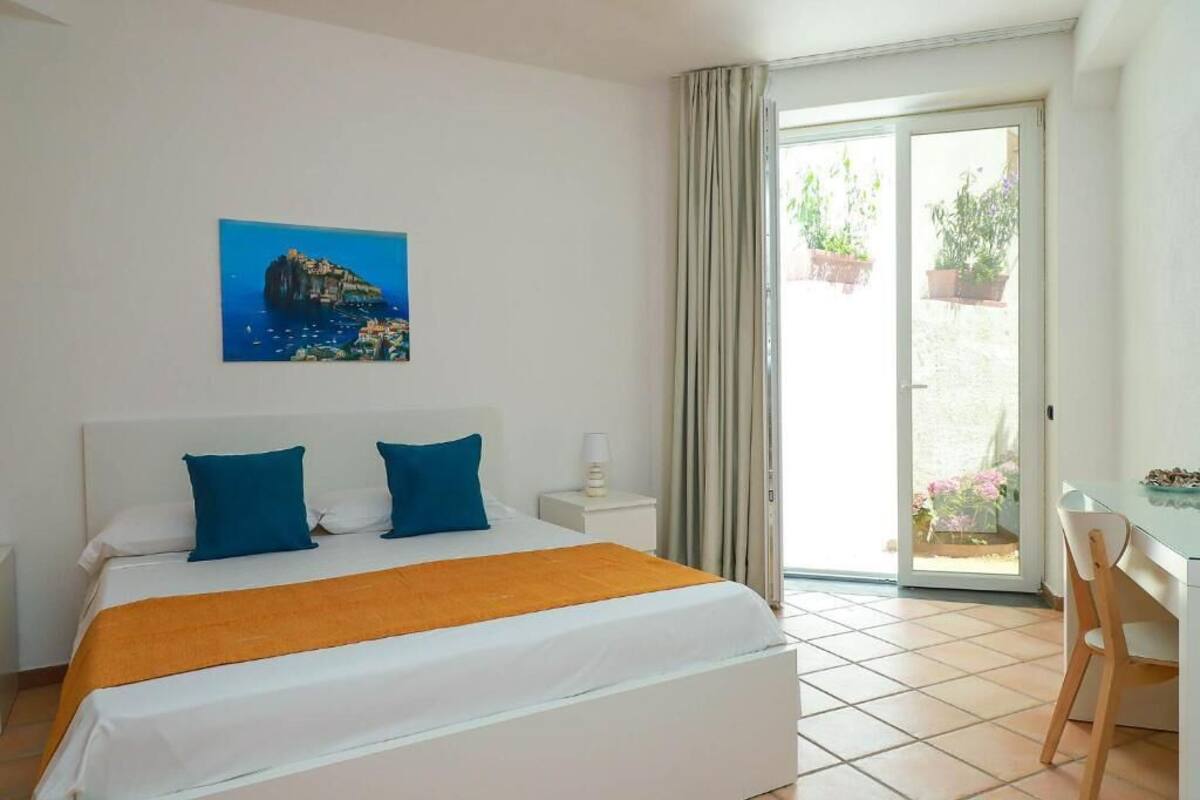 Habitación de hotel con cama, escritorio, silla, mesita de noche, lámpara, un cuadro y una puerta corrediza de cristal que da a un balcón con plantas.