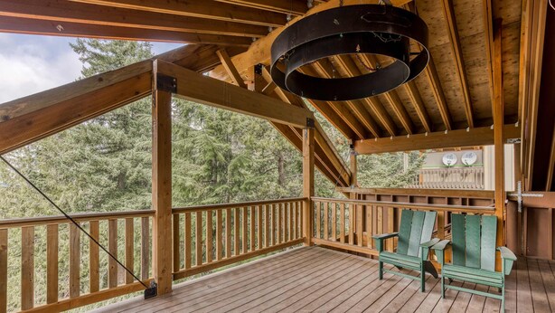 A wooden deck with a view of trees, a hanging light fixture, and two green Adirondack chairs. - Recently updated in 2024-Large, dog-friendly home with covered deck (Government Camp)
