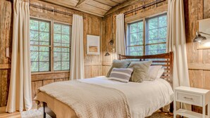Un dormitorio de cabaña de madera con cama, cortinas, una lámpara de noche y una ventana.