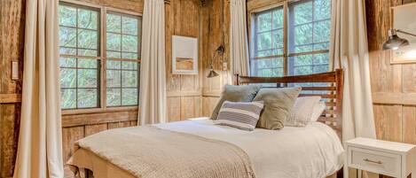 A wooden cabin bedroom with a bed, curtains, a bedside lamp, and a window.