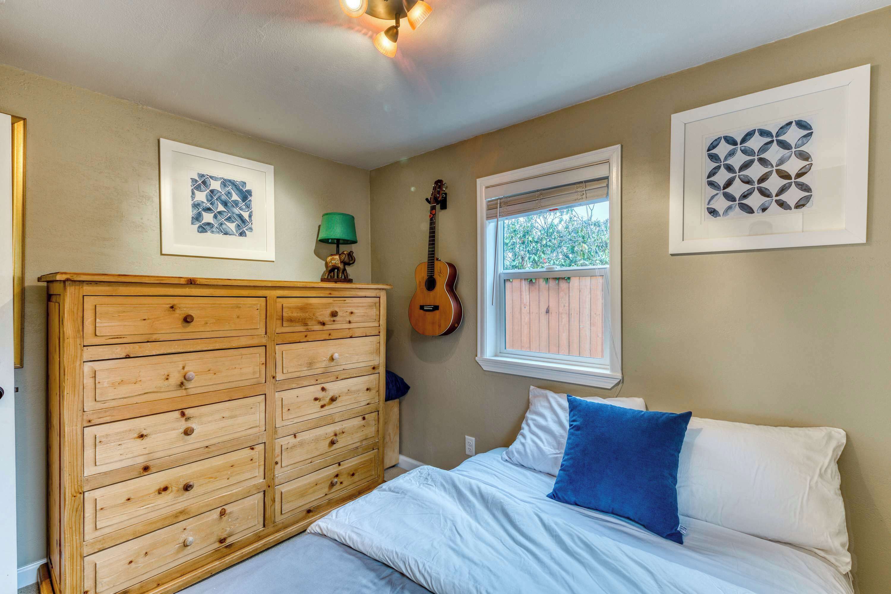 A bedroom with a wooden dresser, a guitar, framed artwork, and a window with blinds.