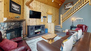 A cozy cabin interior with a stone fireplace, leather furniture, a mounted TV, and a wooden staircase.