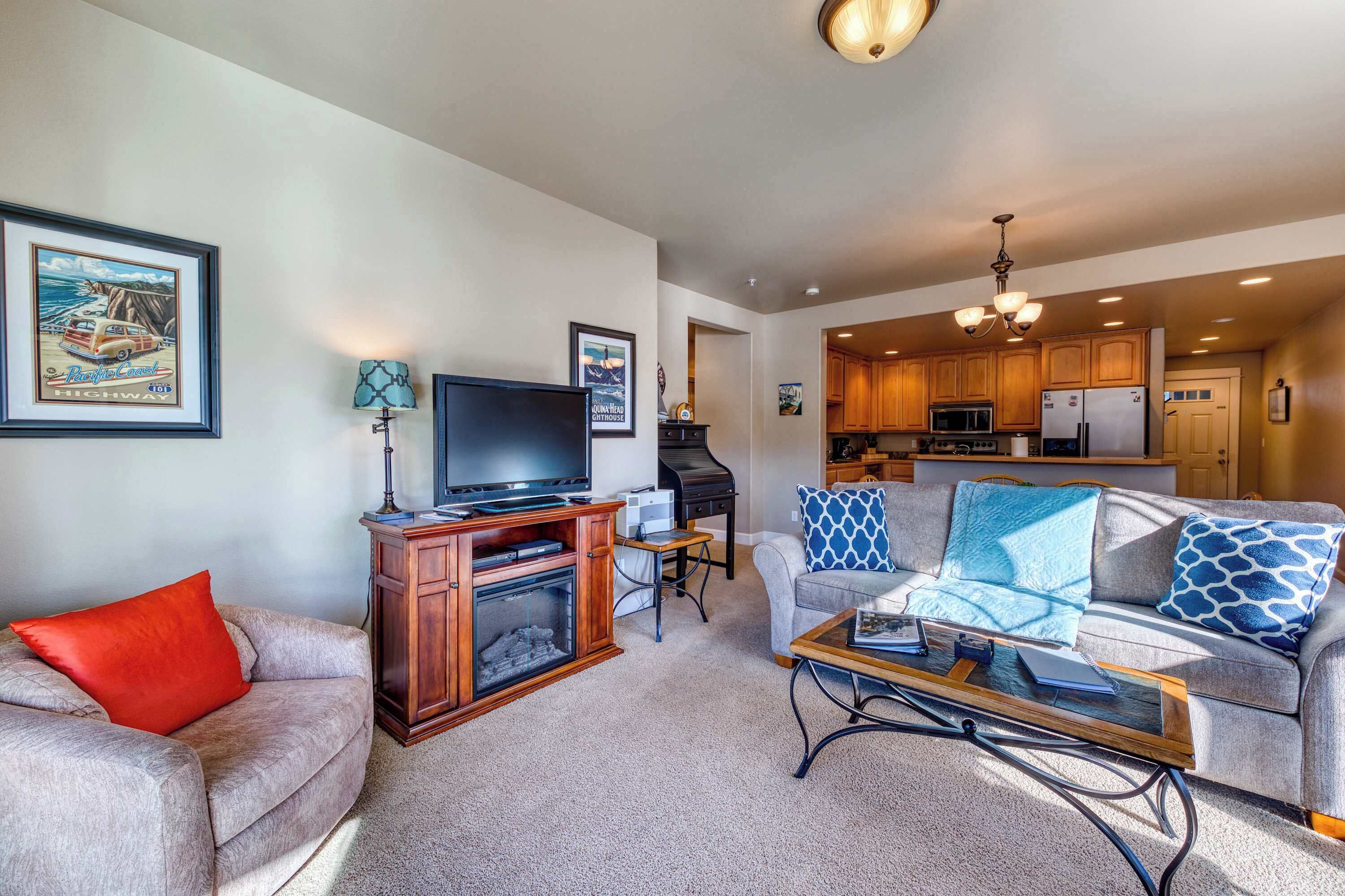 A living room with a sofa, a coffee table, a TV, and a fireplace. There is a kitchen visible in the background.