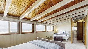 Een slaapkamer met een groot bed, een kleiner bed, een bureau en een stoel. Er zijn meerdere ramen langs de muur en een deur naar een andere kamer.