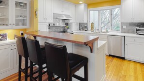 A modern kitchen with white cabinets, a wooden island with bar stools, and a large window providing natural light.