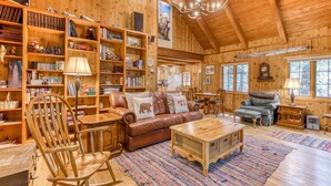 A cozy wooden interior with a leather sofa, bookshelves, and a dining area. - Historic Waterfront Cabin with Hot Tub & Salmon River Access - Dog-Friendly (Welches)