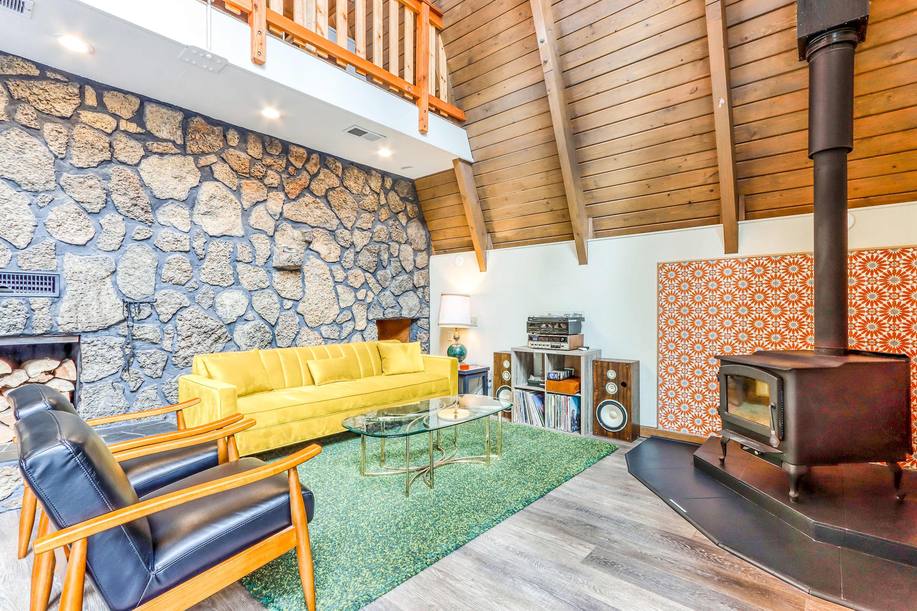 A living room with a stone wall, a yellow sofa, a black leather chair, a glass coffee table, a fireplace, a wooden ceiling, and a wood-burning stove.