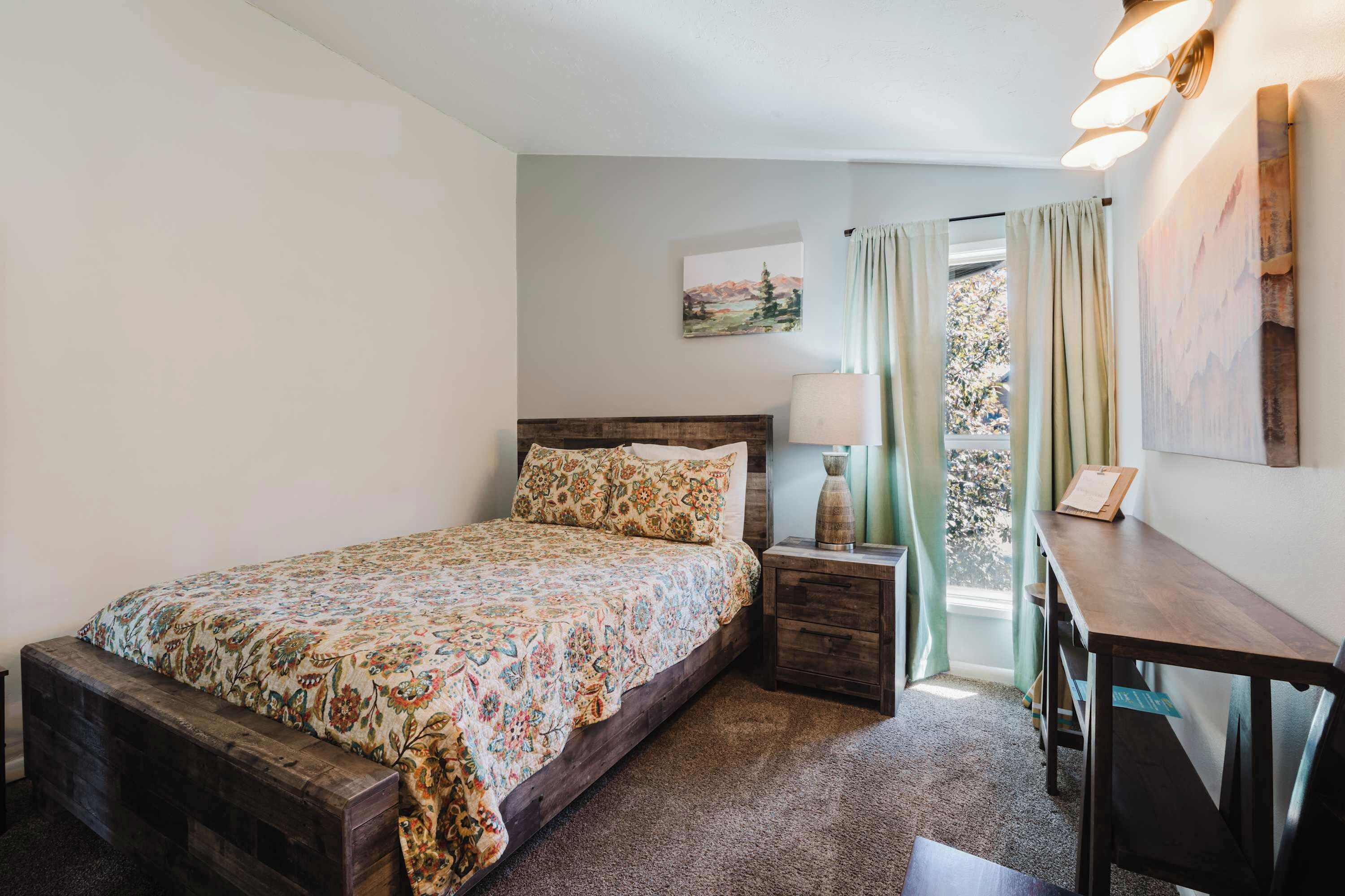 A bedroom with a bed, a desk, a lamp, a window with curtains, and a painting on the wall.