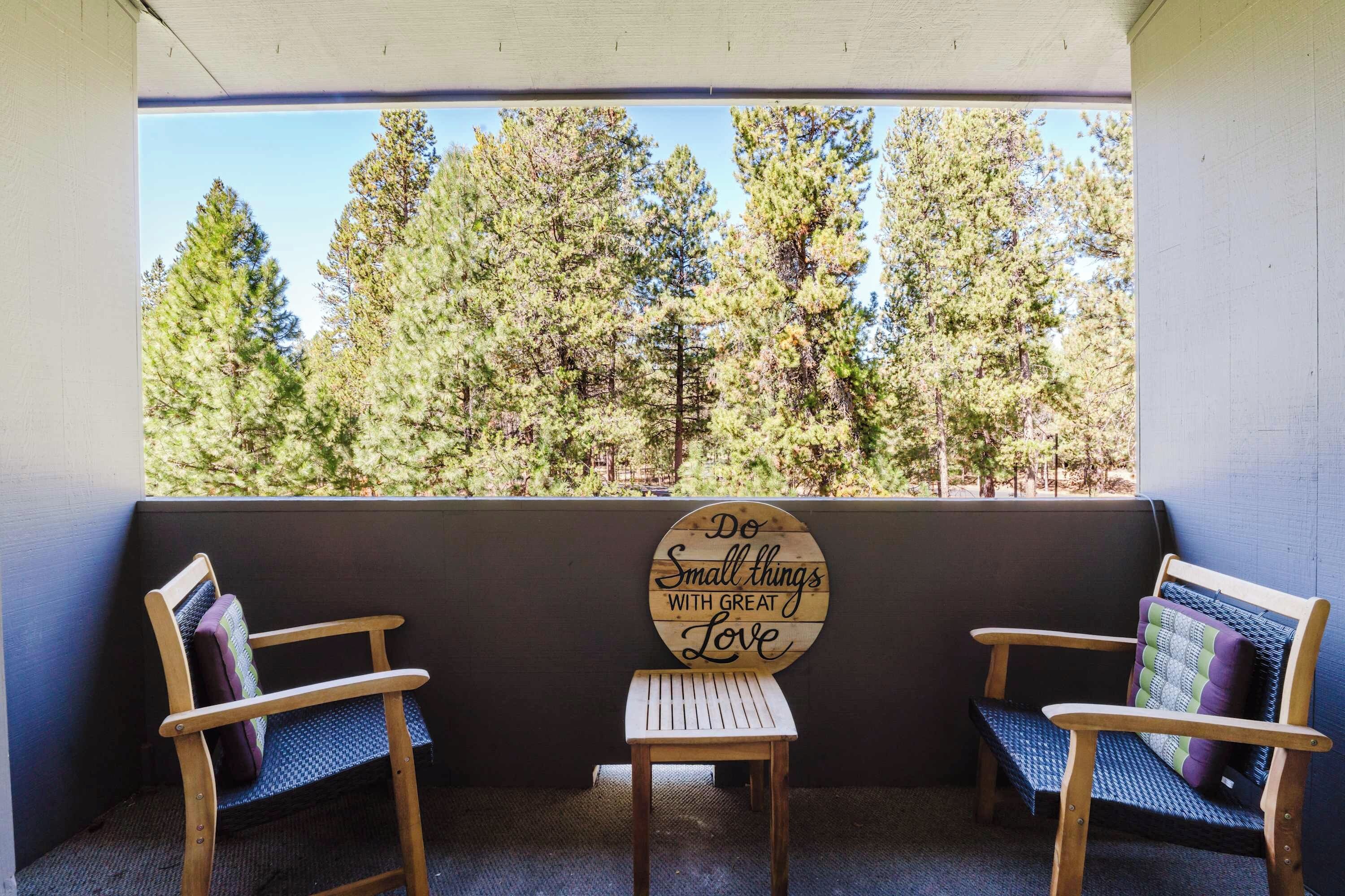 A porch with two wooden chairs and a small table, overlooking a forest. A sign on the wall reads 'Do Small things with Great Love.'
