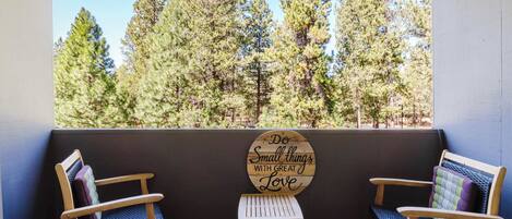 A porch with two wooden chairs and a small table, overlooking a forest. A sign on the wall reads 'Do Small things with Great Love.'