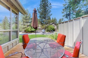Une terrasse couverte avec une table ronde en métal et deux chaises orange, un parasol marron, et une vue sur une rue résidentielle et des arbres.
