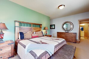 Une chambre à coucher comprenant un lit, des tables de chevet, une commode, un miroir et une porte donnant sur une autre pièce.