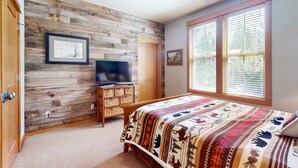 Ein Schlafzimmer mit einer Holzwand, einem Bett mit einem gemusterten Überwurf, einem Fernseher auf einer Holzkommode und einem Fenster mit Jalousien.