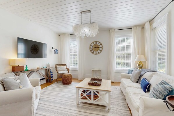 A living room with a flat-screen TV, a clock, a chandelier, a sofa, a coffee table, and a wicker chair. - Stunning Lighthouse Replica Home with Ocean Views - Steps from the Beach (Newport)
